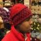 A person wearing a Checkerboard Hat by Halcyon Yarn, featuring a red and navy blue knit design with a braided trim, stands in a yarn shop. Shelves filled with various colorful yarns are visible in the background, while they also don a warm red jacket.