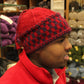 A person wearing a Checkerboard Hat from Halcyon Yarn and a red jacket is seen standing in a shop filled with various colors of yarn on shelves behind them, the warm ambiance enhanced by the soft textures surrounding them.
