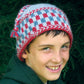 A smiling child wearing the Checkerboard Hat from Halcyon Yarn, featuring red, white, and blue patterns and adorned with a braided trim, stands against a backdrop of green foliage. The child has short brown hair and is dressed in a warm, dark green hoodie.