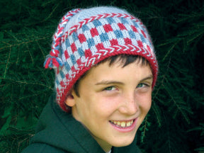 A smiling child wearing the Checkerboard Hat from Halcyon Yarn, featuring red, white, and blue patterns and adorned with a braided trim, stands against a backdrop of green foliage. The child has short brown hair and is dressed in a warm, dark green hoodie.