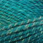 A close-up image of a skein of Plymouth Encore Worsted Yarn from Plymouth Yarn Co. in varying shades of blue and green. The worsted weight yarn is tightly wound into an oblong shape, showcasing its soft and textured fibers. The background is plain and white, highlighting the yarn's colors and texture.