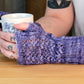 A person with tattoos on their arms is wearing Cupid's Victim Mitts by Halcyon Yarn in a crocheted purple design featuring a delicate Little Arrowhead lace pattern, and holding a white mug adorned with blue floral designs. The background is blurred, focusing on the person's hands and the mug.