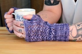 A person with tattoos on their arms is wearing Cupid's Victim Mitts by Halcyon Yarn in a crocheted purple design featuring a delicate Little Arrowhead lace pattern, and holding a white mug adorned with blue floral designs. The background is blurred, focusing on the person's hands and the mug.