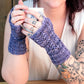 A person with tattooed arms is holding a ceramic mug with both hands. They are wearing Cupid's Victim Mitts, purple, hand-knitted fingerless mitts made from Silky Merino yarn by Halcyon Yarn, featuring a Little Arrowhead lace pattern. The person is dressed in a sleeveless white top, suggesting a cozy and relaxed atmosphere.