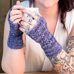 A person with tattoos on their arms is wearing lavender-colored Cupid's Victim Mitts from Halcyon Yarn and holding a cup. They are seated at a wooden table, and part of their white top is visible. The mitts feature a knitted pattern and ribbed cuffs, showcasing Little Arrowhead lace.