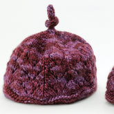 A luxurious Halcyon Yarn's Luxury Lace Noggin Topper in shades of burgundy, purple, and pink, featuring a textured pattern and a small knotted loop at the top. The hat is positioned against a neutral background.