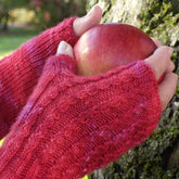 Close-up of hands wearing Halcyon Yarn's Endless Ruby Mitts, holding a red apple against the backdrop of a moss-covered tree trunk. The person seems to be outdoors in a natural setting, and the knitted texture of the gloves is prominently detailed.