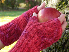 Hands adorned with the red Endless Ruby Mitts by Halcyon Yarn, crafted from the luxurious Silky Merino yarn, are grasping a red apple against the trunk of a tree in an outdoor setting.