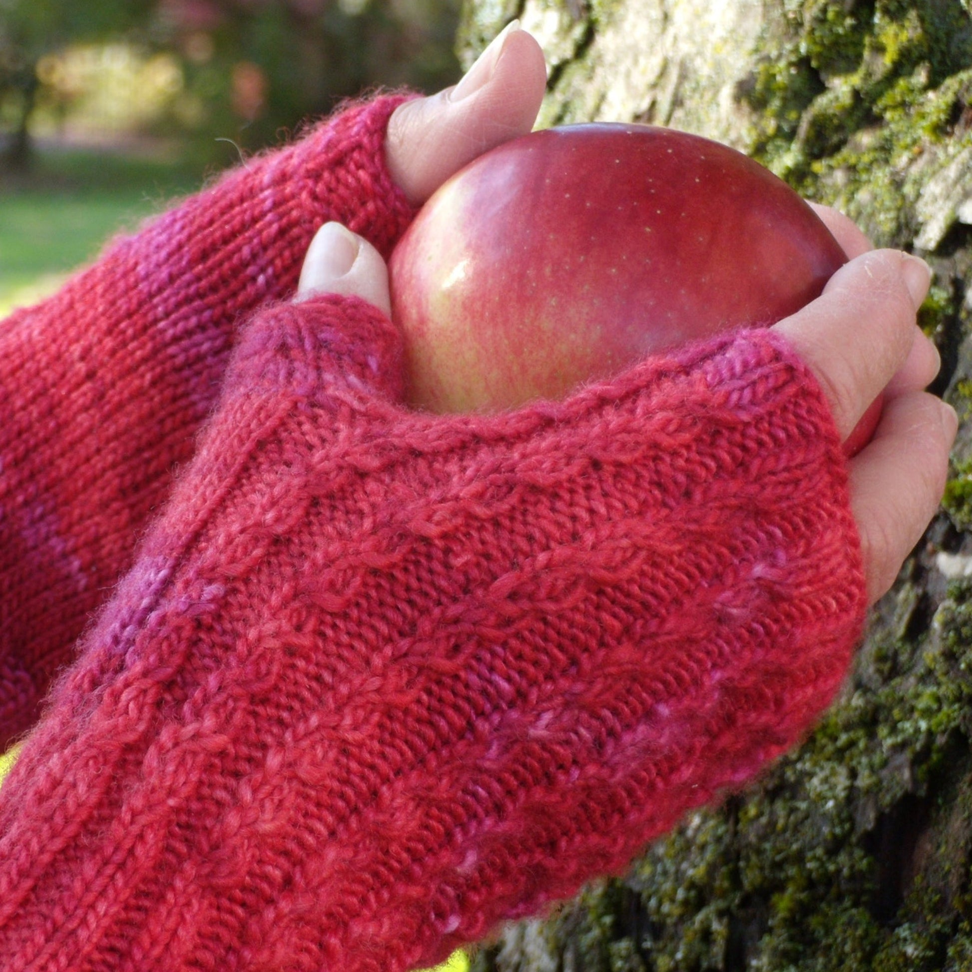 Hands wearing red knitted Endless Ruby Mitts, a downloadable pattern from Halcyon Yarn crafted from silky Merino yarn, holding a red apple near a tree with moss.