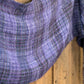 A close-up of a Halcyon Yarn Block After Block Woven Scarf, showcasing its purple and blue hues draped over a wooden surface. The texture of the textiles is visible, highlighting a blend of shades in a checkered weaving pattern against the rustic backdrop provided by the wood.