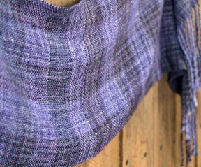 A close-up of a Halcyon Yarn Block After Block Woven Scarf, showcasing its purple and blue hues draped over a wooden surface. The texture of the textiles is visible, highlighting a blend of shades in a checkered weaving pattern against the rustic backdrop provided by the wood.