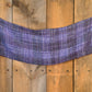 A purple plaid scarf with fringed ends is draped horizontally against a wooden backdrop. The Block After Block Woven Scarf from Halcyon Yarn features various shades of purple and blue with a woven texture, adding contrast to the rustic wooden background.