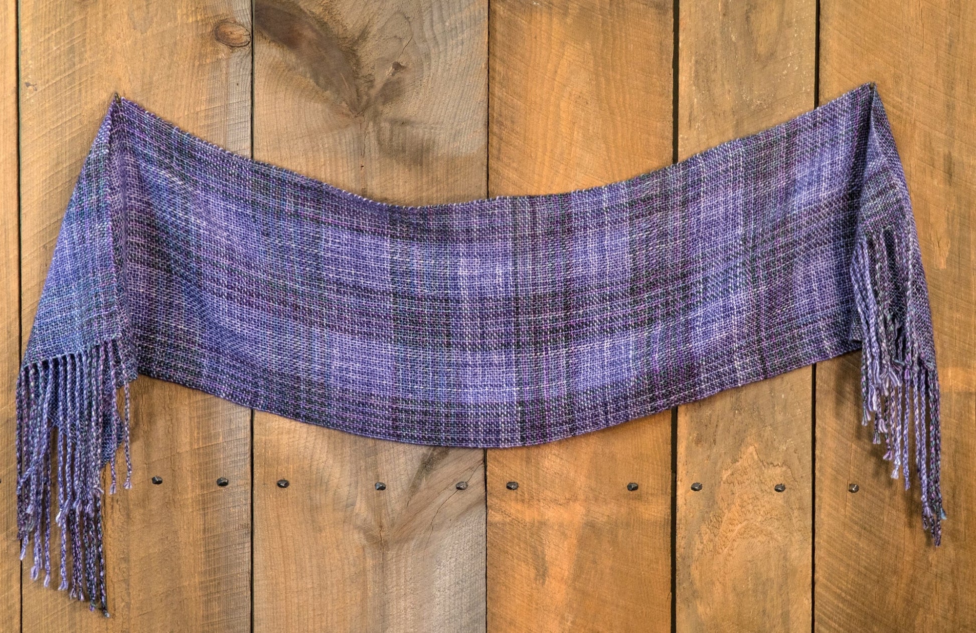 A purple plaid scarf with fringed ends is draped horizontally against a wooden backdrop. The Block After Block Woven Scarf from Halcyon Yarn features various shades of purple and blue with a woven texture, adding contrast to the rustic wooden background.