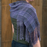 A person wearing a green shirt and blue jeans models the "Block After Block Woven Scarf" from Halcyon Yarn, featuring a handwoven plaid pattern in purple and blue hues. Made with Malabrigo Silky Merino yarn in kettle-dyed colors, the scarf is adorned with long fringes hanging from the edge. The background consists of wooden panels.