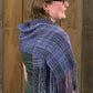 A person with short hair and glasses is wearing a green top and the Block After Block Woven Scarf by Halcyon Yarn, a fringed purple scarf featuring an intricate weaving pattern, draped over their shoulders. They are looking to the right, standing in front of a wooden background.