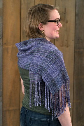 A person with short hair and glasses is wearing a green top and the Block After Block Woven Scarf by Halcyon Yarn, a fringed purple scarf featuring an intricate weaving pattern, draped over their shoulders. They are looking to the right, standing in front of a wooden background.