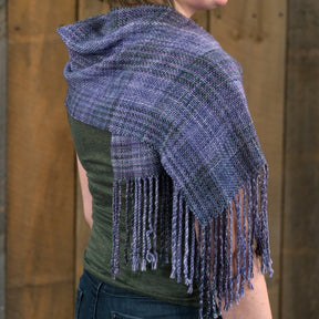 A person is wearing a handmade, woven Block After Block Woven Scarf in a plaid pattern with shades of purple and green. The Halcyon Yarn scarf features long, twisted fringe along the edge. The person is turned away, showcasing the intricate weaving pattern against a wooden backdrop.