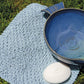 A blue Textured Cotton Washcloth from Halcyon Yarn, featuring a slip stitch pattern, is spread on the grass. On it rests a textured blue and white pottery bowl filled with water and a white soap bar imprinted with a dove design. The outdoor setting suggests a natural and fresh ambiance.