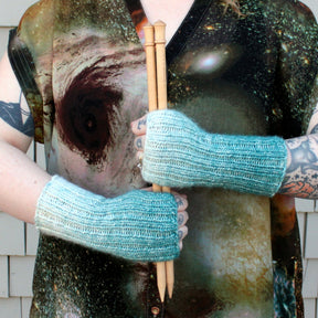 A person wearing a space-themed shirt holds four wooden knitting needles with both hands. They are also wearing "Eight Days A Week, Fingerless Mitts" by Halcyon Yarn, perfect for easy mitts projects, with tattoos visible on their arms.