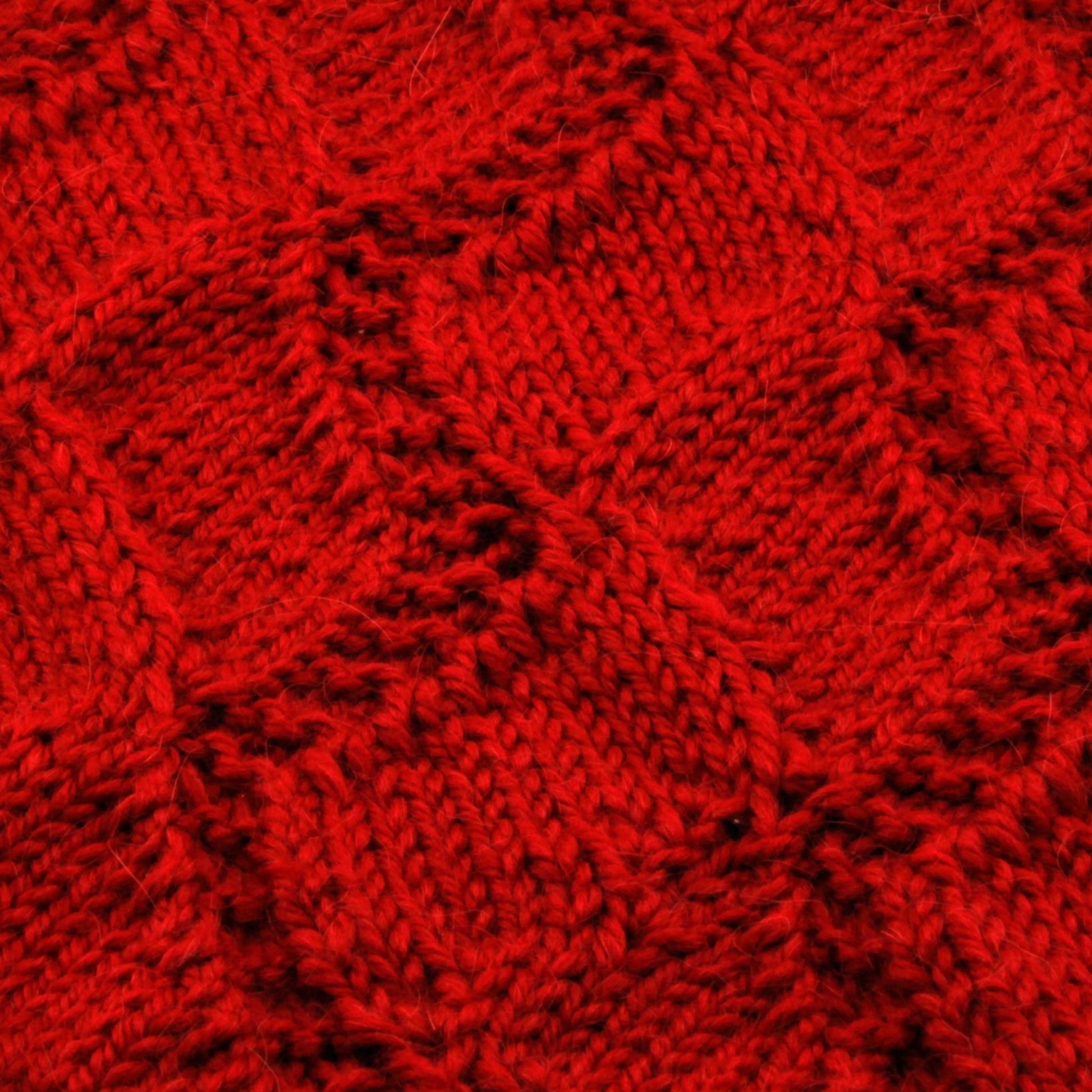 Close-up of the Peruby Waves Throw from Halcyon Yarn, showcasing its red knitted fabric with a diamond pattern. The texture emphasizes intricate detailing and various knitting techniques, including a wave stitch pattern, creating a visually rich and tactile surface.