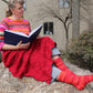 A woman sits on a rock outdoors, reading a large blue book. She wears the Peruby Waves Throw from Halcyon Yarn—a colorful, striped sweater made of chunky yarn—paired with a red knitted skirt and matching striped socks in shades of orange, pink, and red. Behind her are bushes and a beige building with bare trees—a scene of peaceful handmade beauty.