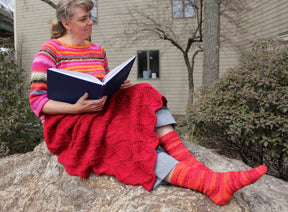 A woman sits on a rock outdoors, reading a large blue book. She wears the Peruby Waves Throw from Halcyon Yarn—a colorful, striped sweater made of chunky yarn—paired with a red knitted skirt and matching striped socks in shades of orange, pink, and red. Behind her are bushes and a beige building with bare trees—a scene of peaceful handmade beauty.