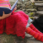 A person dressed in a colorful knitted sweater and socks is sitting outdoors on a stone bench, reading a book. The Peruby Waves Throw from Halcyon Yarn, crafted with chunky yarn, is draped over their lap. The background features stacked stones, adding to the cozy outdoor setting and highlighting the handmade beauty of the scene.