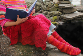 A person dressed in a colorful knitted sweater and socks is sitting outdoors on a stone bench, reading a book. The Peruby Waves Throw from Halcyon Yarn, crafted with chunky yarn, is draped over their lap. The background features stacked stones, adding to the cozy outdoor setting and highlighting the handmade beauty of the scene.