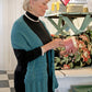 A woman with short blonde hair, wearing the Gillies Wrap from Halcyon Yarn—a teal knit shawl adorned with delicate lace motifs—over a black turtleneck, is holding and examining a pink candle jar in a quaint shop. The shawl complements the colorful items and decorative pieces displayed around her. An Adobe PDF icon is overlaid at the top of the image.