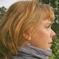 The product image features a woman with light brown hair shown in profile, facing to the right. She is wearing the Reversible Cowl from Halcyon Yarn, a knitted gray scarf perfect for keeping warm in cold weather, and dangling earrings. The background includes lush greenery, suggesting an outdoor setting with natural light.