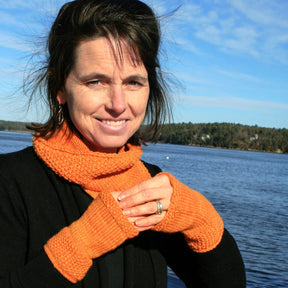 A woman stands outdoors by a body of water, framed by a tree-lined shore and blue sky. She is wearing a black sweater and the "Simply Beautiful Cowl & Mitts" set from Halcyon Yarn, featuring an orange seed stitch cowl and matching orange fingerless gloves crafted from worsted yarn. Smiling at the camera, her hands are clasped together in contentment.