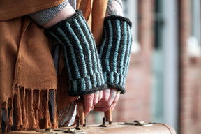 A person wears Corrugated Mitts, fingerless gloves in blue and dark gray from Halcyon Yarn, reminiscent of dapper mitts, gripping the handle of a vintage suitcase. They are dressed in a brown shawl, standing by a brick wall. Their hands are visible with some tattoos on their wrist.