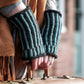 A person wearing Corrugated Mitts from Halcyon Yarn, characterized by their brown and blue stripes, holds a vintage suitcase. The individual is dressed in a brown fringed jacket, while the blurred background reveals a brick wall and windows.