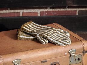 A pair of Corrugated Mitts from Halcyon Yarn, featuring white and olive green stripes, lies on top of a vintage brown suitcase. The suitcase, adorned with metal latches and displaying signs of wear that hint at its age, stands out against a background with a brick wall and dark wooden accents.