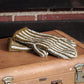 The "Corrugated Mitts, download" by Halcyon Yarn, featuring stylish white and olive green stripes with intricate colorwork knitting, are elegantly displayed on a closed, weathered suitcase with metal clasps. A wooden bench behind the suitcase is set against a brick wall backdrop.