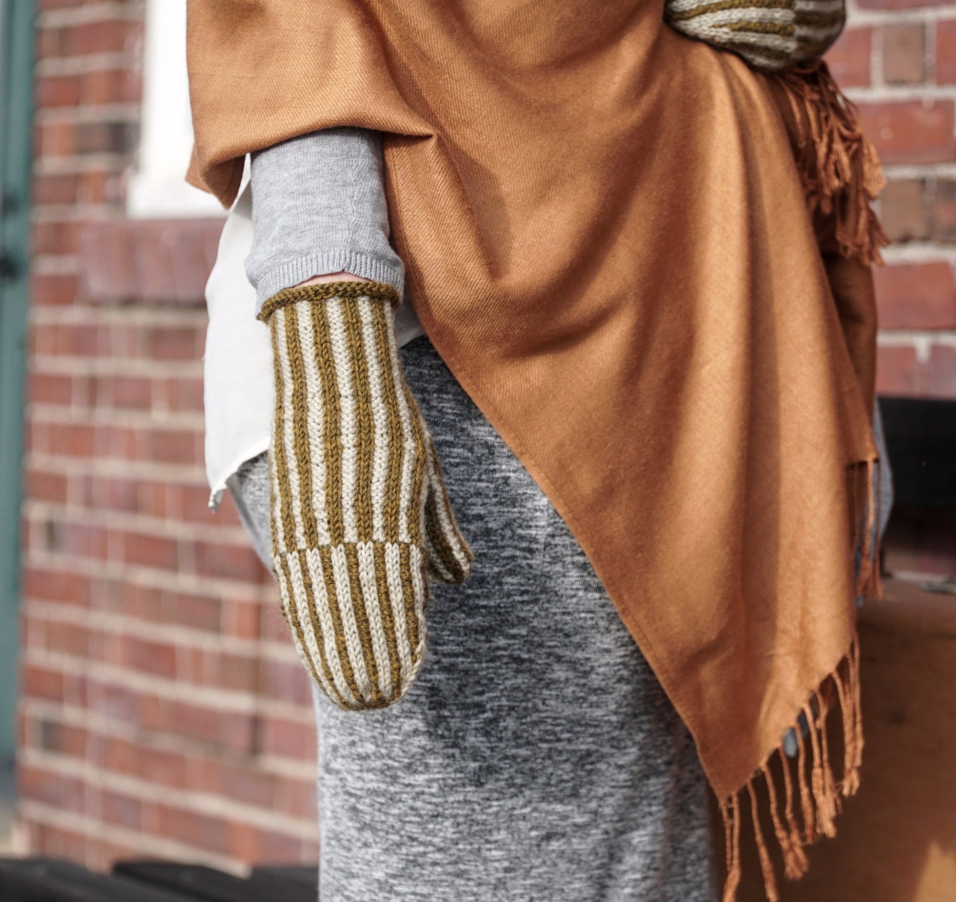 A person is standing outside in front of a brick wall, dressed warmly in grey and white clothing, a brown shawl, and brown-and-white striped mittens from Halcyon Yarn's Corrugated Mitts collection. The person appears to be holding a brown bag or package.