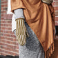 A person is draped in a brown shawl and wears a gray outfit. They are also wearing Halcyon Yarn's Corrugated Mitts, featuring green and white stripes. They stand in front of a brick wall.