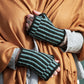 A person, adorned in a mustard-colored shawl and a gray sweater, showcases the dapper Corrugated Mitts by Halcyon Yarn. These mitts feature exquisite blue and black stripes in intricate colorwork knitting, while tattoos embellish their hand and fingers as they grasp the shawl closed around their neck.