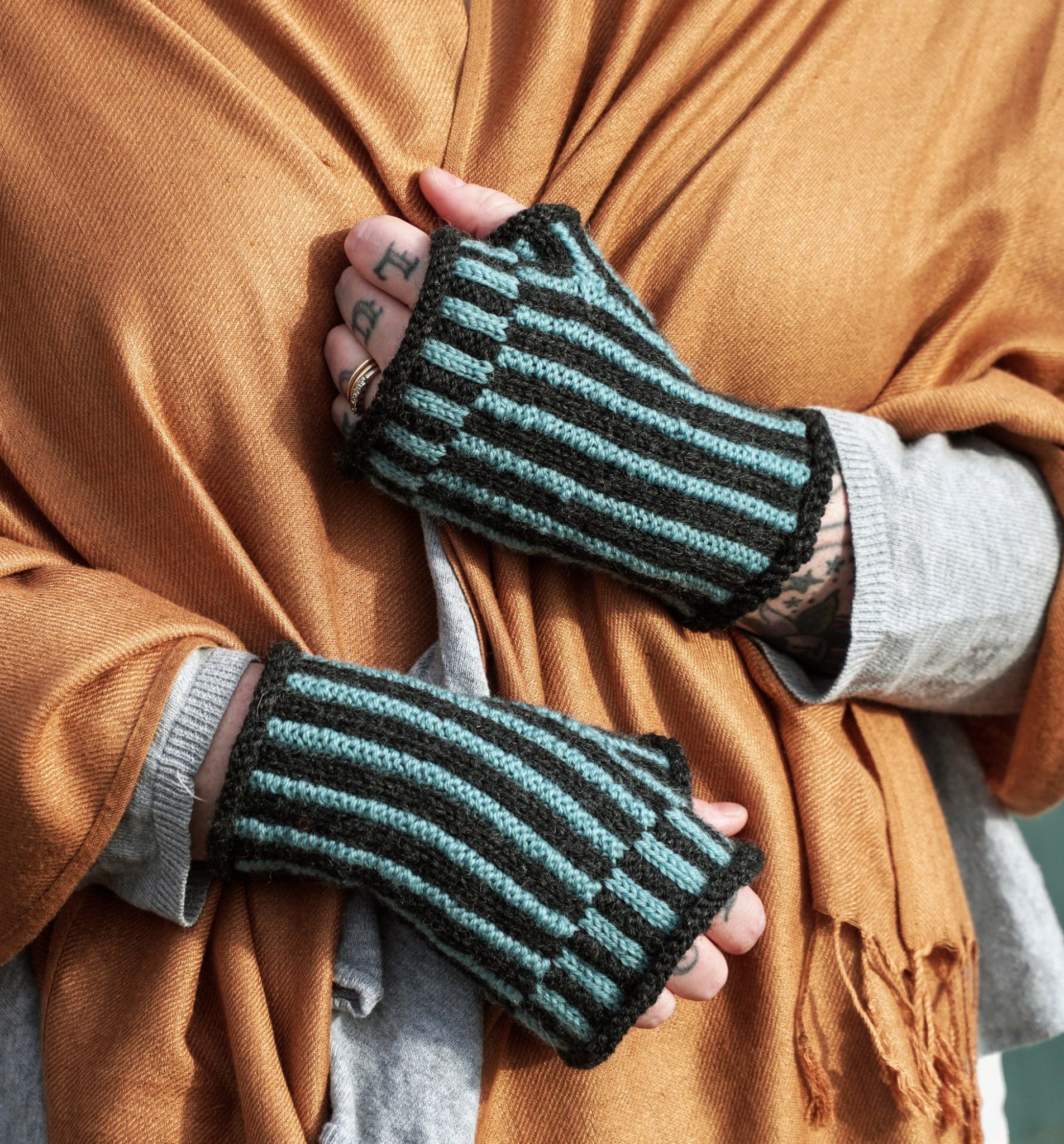 A person, adorned in a mustard-colored shawl and a gray sweater, showcases the dapper Corrugated Mitts by Halcyon Yarn. These mitts feature exquisite blue and black stripes in intricate colorwork knitting, while tattoos embellish their hand and fingers as they grasp the shawl closed around their neck.