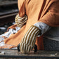 A person wearing a cozy orange shawl and Halcyon Yarn's Corrugated Mitts, which feature intricate colorwork knitting, rests their hands on a vintage suitcase. The background is slightly blurred, emphasizing the person's attire and the suitcase. Rails on a train track are visible, suggesting a travel or departure theme.