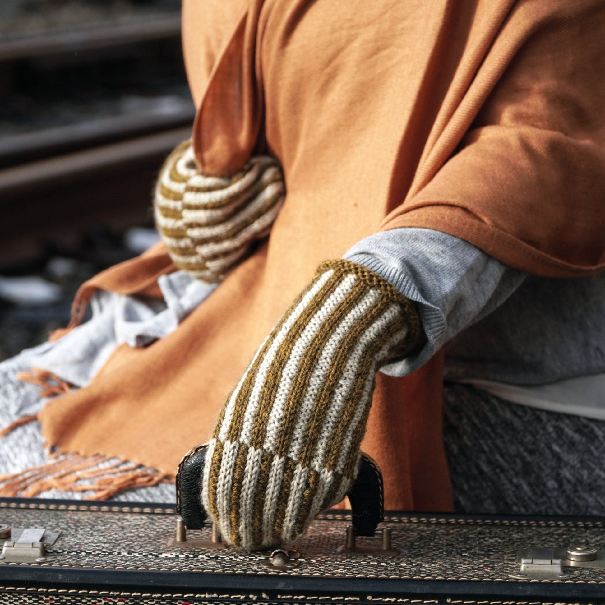 A person wearing a cozy orange shawl and Halcyon Yarn's Corrugated Mitts, which feature intricate colorwork knitting, rests their hands on a vintage suitcase. The background is slightly blurred, emphasizing the person's attire and the suitcase. Rails on a train track are visible, suggesting a travel or departure theme.