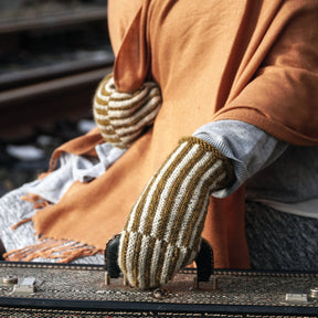 A person wearing Corrugated Mitts by Halcyon Yarn, featuring beige and white stripes, grips the handle of a vintage suitcase. The individual is dressed in a light gray top and an orange shawl with fringes, standing near train tracks.