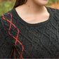 A woman is wearing a dark grey Halcyon Yarn Hearthstone Cabled Pullover with a red braided pattern on one shoulder. The background is a blurred outdoor scene with autumn leaves on the ground.