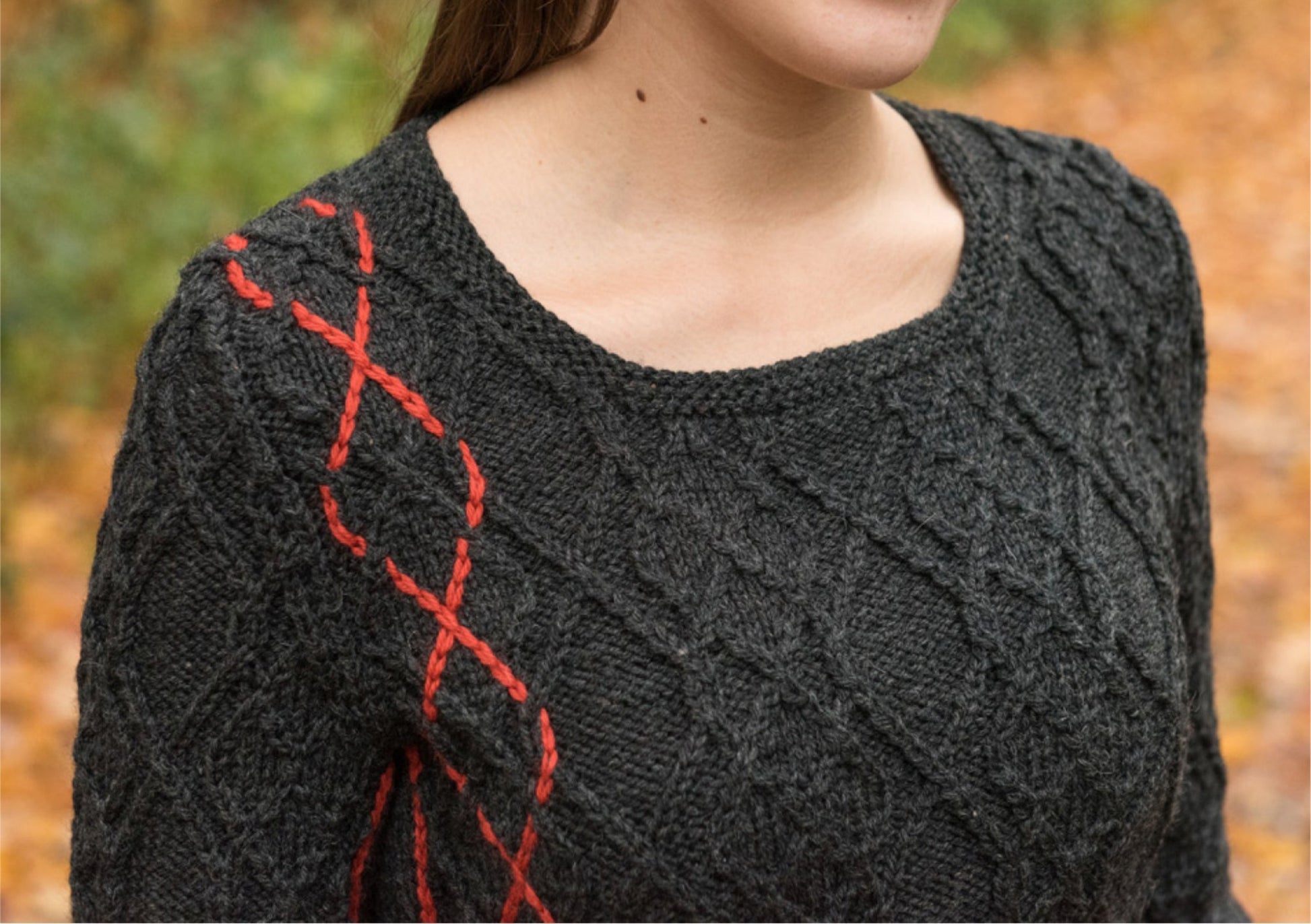 A woman is wearing a dark grey Halcyon Yarn Hearthstone Cabled Pullover with a red braided pattern on one shoulder. The background is a blurred outdoor scene with autumn leaves on the ground.