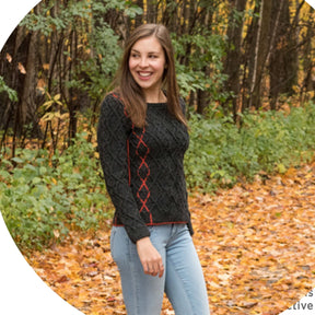 A woman with long brown hair is smiling while walking on a leaf-covered path in a forest. She is wearing a Halcyon Yarn's Hearthstone Cabled Pullover paired with light blue jeans. The trees in the background are filled with green and yellow leaves.