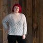 A person with short, vibrant pink hair is standing against a wooden background. They are wearing the Halcyon Yarn Atmosphere Lace Mohair Pullover, a light, vintage-inspired sweater featuring a delicate lace pattern, paired with black pants. With one hand resting on their hip and a neutral expression on their face, they present an effortlessly stylish look.