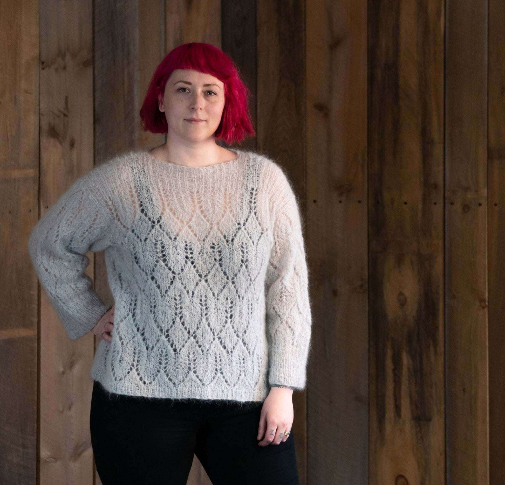 A person with short, vibrant pink hair is standing against a wooden background. They are wearing the Halcyon Yarn Atmosphere Lace Mohair Pullover, a light, vintage-inspired sweater featuring a delicate lace pattern, paired with black pants. With one hand resting on their hip and a neutral expression on their face, they present an effortlessly stylish look.