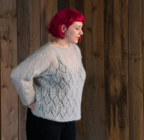 A person with short, bright pink hair stands in profile against a wooden wall. They wear a loose, cream-colored Halcyon Yarn Atmosphere Lace Mohair Pullover with a subtle diamond lace pattern and black pants. Their hands rest on their hips as they look to the side.