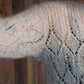 Close-up of a person's arm wearing an Atmosphere Lace Mohair Pullover by Halcyon Yarn in light grey, featuring an intricately knitted diamond lace pattern. The background is a rustic wooden wall, enhancing the vintage-inspired charm of the pullover.