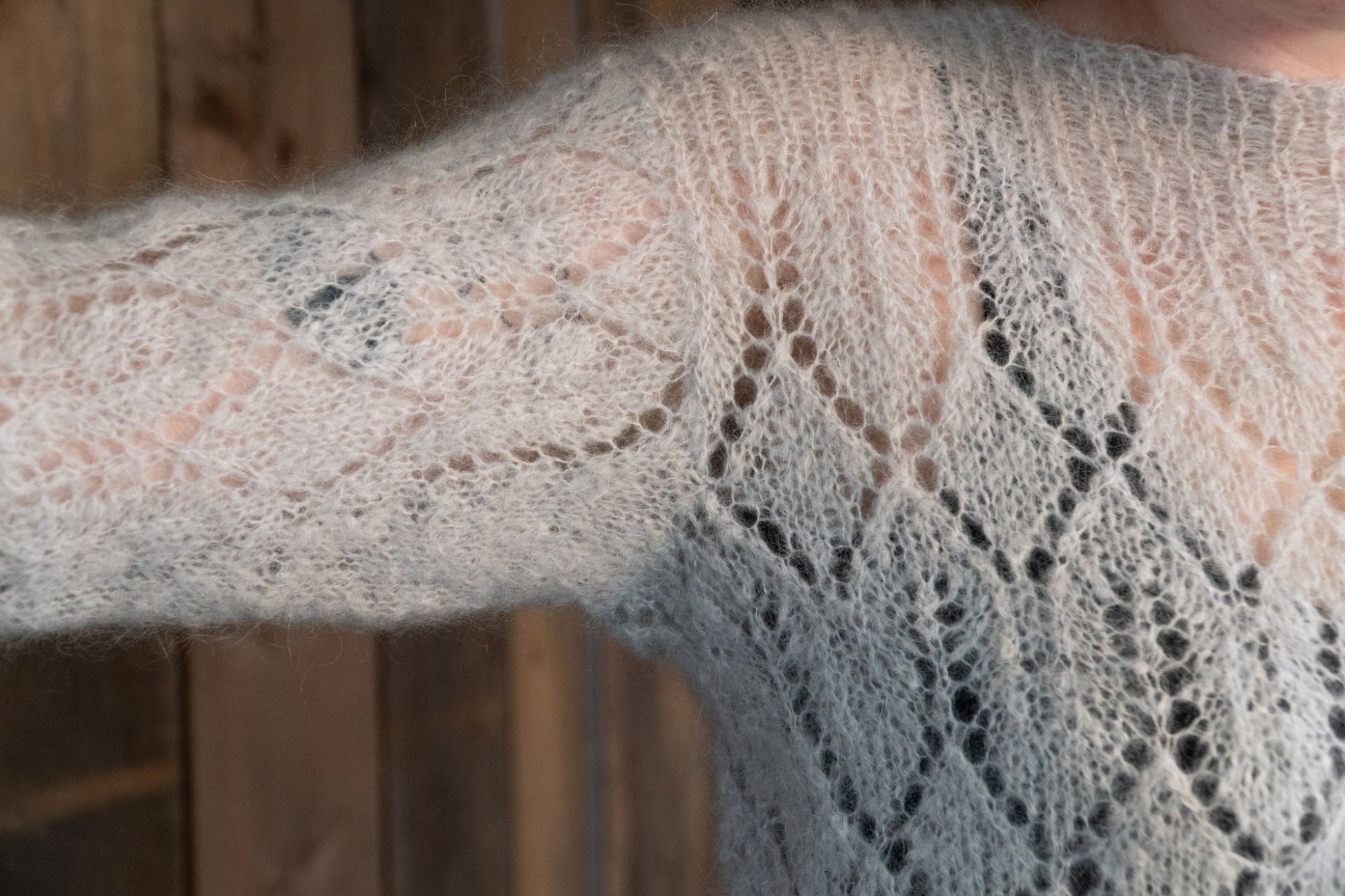 Close-up of a person's arm wearing an Atmosphere Lace Mohair Pullover by Halcyon Yarn in light grey, featuring an intricately knitted diamond lace pattern. The background is a rustic wooden wall, enhancing the vintage-inspired charm of the pullover.